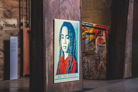 A stylized portrait of a person with long hair is displayed on a wooden panel in an urban setting. The artwork features a color scheme of red, blue, and beige with a bold message at the bottom reading 'We The People - Protect Each Other'. The background includes graffiti on metal structures and industrial elements, giving a gritty, artistic atmosphere.
