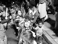 A group of children and adults gather on a street, engaging in playful activities. The children exhibit joyful expressions, with one girl reaching upward, suggesting a festive and lively atmosphere. The scene is crowded, indicating a public event or gathering.