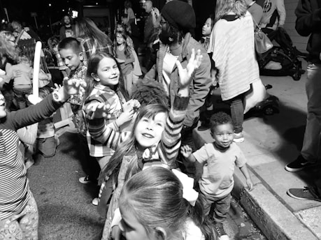 A group of children and adults gather on a street, engaging in playful activities. The children exhibit joyful expressions, with one girl reaching upward, suggesting a festive and lively atmosphere. The scene is crowded, indicating a public event or gathering.