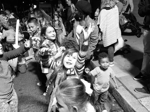 A group of children and adults gather on a street, engaging in playful activities. The children exhibit joyful expressions, with one girl reaching upward, suggesting a festive and lively atmosphere. The scene is crowded, indicating a public event or gathering.