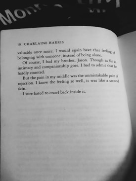 A black and white photograph of an open book. The visible page contains text, including the author's name 'Charlaine Harris' and part of a narrative about feelings of loneliness and rejection. A portion of black fabric with white lettering is partially visible at the top.