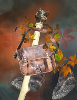 A rustic leather satchel is suspended on a carved wooden staff, with autumn leaves artistically arranged around it. Below the satchel, a pair of worn leather boots are positioned, adding to the rugged outdoor theme. The background blends warm earthy tones with hints of dark green and orange.
