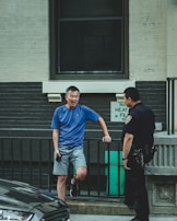 A man in a blue t-shirt and shorts is standing casually next to a black railing, leaning slightly on one leg while holding a phone. He is engaged in conversation with a police officer wearing a dark uniform. The scene is set in an urban environment with a gray and white brick wall in the background.