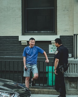 A man in a blue t-shirt and shorts is standing casually next to a black railing, leaning slightly on one leg while holding a phone. He is engaged in conversation with a police officer wearing a dark uniform. The scene is set in an urban environment with a gray and white brick wall in the background.