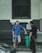A man in a blue t-shirt and shorts is standing casually next to a black railing, leaning slightly on one leg while holding a phone. He is engaged in conversation with a police officer wearing a dark uniform. The scene is set in an urban environment with a gray and white brick wall in the background.