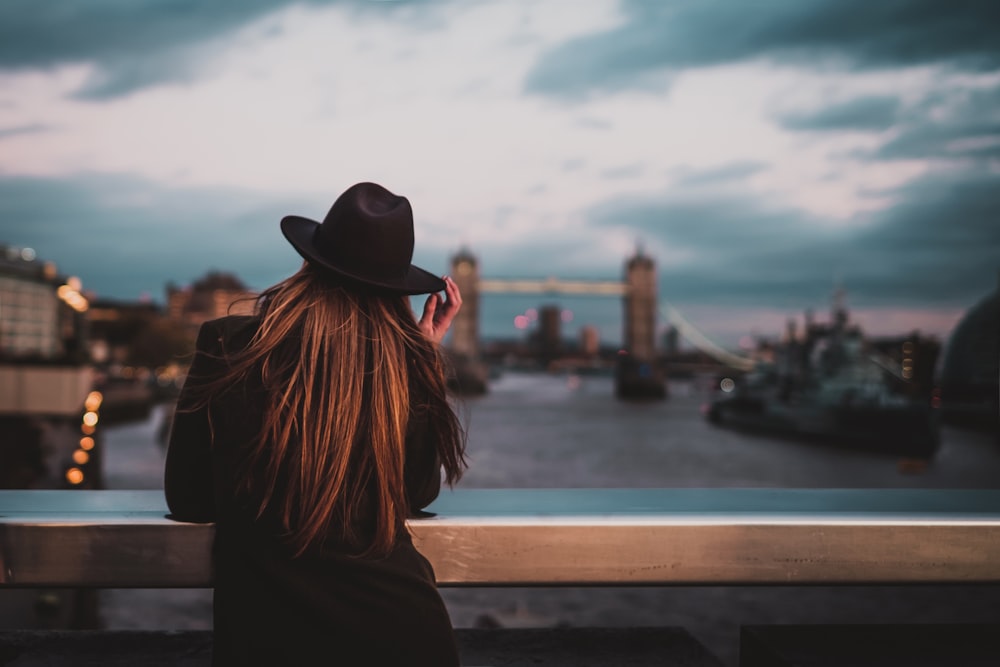 Selektives Fokusfoto des Rückens einer Frau, die einen Fedora-Hut mit Ellbogen auf dem Geländer mit Blick auf die Brücke trägt