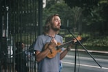 A man with long hair and a beard sings into a microphone while playing a small string instrument, possibly a ukulele, on a street. He wears a striped shirt and appears focused on his performance. In the background, there is greenery and a fenced area, suggesting an outdoor setting.