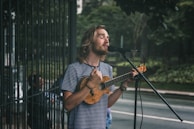 A man with long hair and a beard sings into a microphone while playing a small string instrument, possibly a ukulele, on a street. He wears a striped shirt and appears focused on his performance. In the background, there is greenery and a fenced area, suggesting an outdoor setting.