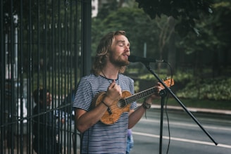 A man with long hair and a beard sings into a microphone while playing a small string instrument, possibly a ukulele, on a street. He wears a striped shirt and appears focused on his performance. In the background, there is greenery and a fenced area, suggesting an outdoor setting.