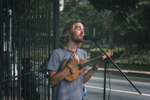 A man with long hair and a beard sings into a microphone while playing a small string instrument, possibly a ukulele, on a street. He wears a striped shirt and appears focused on his performance. In the background, there is greenery and a fenced area, suggesting an outdoor setting.