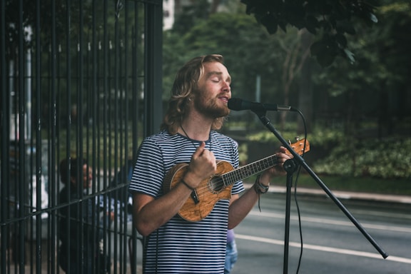 A man with long hair and a beard sings into a microphone while playing a small string instrument, possibly a ukulele, on a street. He wears a striped shirt and appears focused on his performance. In the background, there is greenery and a fenced area, suggesting an outdoor setting.