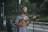 A man with long hair and a beard sings into a microphone while playing a small string instrument, possibly a ukulele, on a street. He wears a striped shirt and appears focused on his performance. In the background, there is greenery and a fenced area, suggesting an outdoor setting.