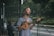 A man with long hair and a beard sings into a microphone while playing a small string instrument, possibly a ukulele, on a street. He wears a striped shirt and appears focused on his performance. In the background, there is greenery and a fenced area, suggesting an outdoor setting.