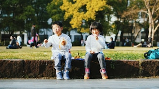 Two children are sitting on a low stone wall in a park, enjoying snacks. The boy on the left is holding and eating a banana, while the girl on the right is holding a juice box. They are both dressed casually and appear to be having fun. In the background, other people are relaxing on the grass under the shade of trees in a sunny, outdoor setting.