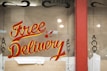 A window display featuring the words 'Free Delivery' painted in bold, red and yellow script. The background is brown with some decorative black designs on the sides. There is a red frame around the window and cardboard packaging material at the bottom.