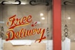 A window display featuring the words 'Free Delivery' painted in bold, red and yellow script. The background is brown with some decorative black designs on the sides. There is a red frame around the window and cardboard packaging material at the bottom.