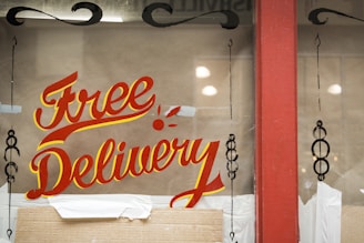 A window display featuring the words 'Free Delivery' painted in bold, red and yellow script. The background is brown with some decorative black designs on the sides. There is a red frame around the window and cardboard packaging material at the bottom.