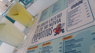 A menu with a distinctive quote and colorful cartoon graphic is prominently featured. Two glasses filled with a light yellow beverage are placed next to the menu. The layout includes sections with lists of food and drinks, indicating a restaurant or cafe setting.