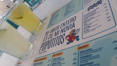A menu with a distinctive quote and colorful cartoon graphic is prominently featured. Two glasses filled with a light yellow beverage are placed next to the menu. The layout includes sections with lists of food and drinks, indicating a restaurant or cafe setting.