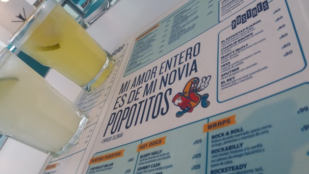 A menu with a distinctive quote and colorful cartoon graphic is prominently featured. Two glasses filled with a light yellow beverage are placed next to the menu. The layout includes sections with lists of food and drinks, indicating a restaurant or cafe setting.