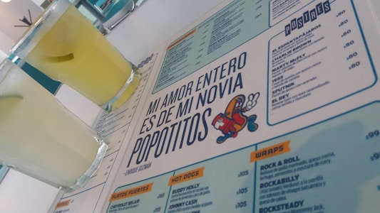 A menu with a distinctive quote and colorful cartoon graphic is prominently featured. Two glasses filled with a light yellow beverage are placed next to the menu. The layout includes sections with lists of food and drinks, indicating a restaurant or cafe setting.