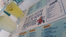 A menu with a distinctive quote and colorful cartoon graphic is prominently featured. Two glasses filled with a light yellow beverage are placed next to the menu. The layout includes sections with lists of food and drinks, indicating a restaurant or cafe setting.