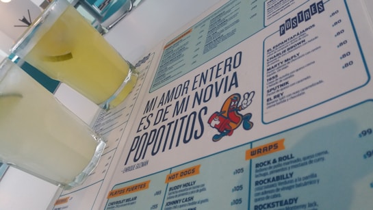 A menu with a distinctive quote and colorful cartoon graphic is prominently featured. Two glasses filled with a light yellow beverage are placed next to the menu. The layout includes sections with lists of food and drinks, indicating a restaurant or cafe setting.