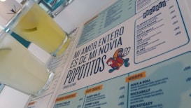 A menu with a distinctive quote and colorful cartoon graphic is prominently featured. Two glasses filled with a light yellow beverage are placed next to the menu. The layout includes sections with lists of food and drinks, indicating a restaurant or cafe setting.