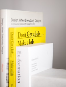 Several books are stacked vertically with their spines facing outward. The titles are visible, including 'Design, When Everybody Designs,' 'Don't Get a Job... Make a Job,' and 'Ex-formation.' The books have a clean and minimalist aesthetic, with prominent use of white and yellow colors.