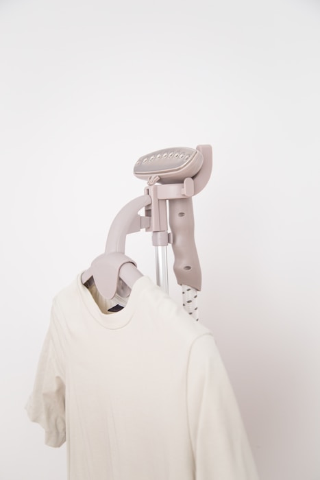 A clothes steamer is placed above a white t-shirt on a hanger. The steamer has a light beige color and features a sleek, modern design. The t-shirt appears to be made of a soft, plain fabric.