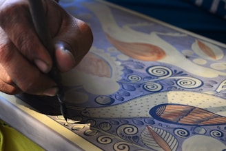 A close-up view of a person's hand holding a tool, meticulously drawing intricate patterns and designs on a surface. The artwork features detailed bird and floral motifs in shades of blue, orange, and cream. The person's focus and precision are evident in the careful strokes.