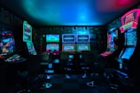Dimly lit gaming arcade featuring a variety of brightly illuminated slot machines and video gaming screens. Neon signs and colorful displays create a vibrant atmosphere. The arrangement of gaming machines suggests an inviting space for entertainment, with chairs positioned for players.