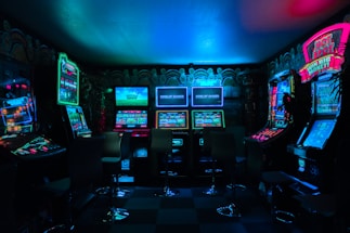 Dimly lit gaming arcade featuring a variety of brightly illuminated slot machines and video gaming screens. Neon signs and colorful displays create a vibrant atmosphere. The arrangement of gaming machines suggests an inviting space for entertainment, with chairs positioned for players.