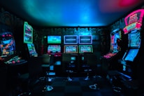 Dimly lit gaming arcade featuring a variety of brightly illuminated slot machines and video gaming screens. Neon signs and colorful displays create a vibrant atmosphere. The arrangement of gaming machines suggests an inviting space for entertainment, with chairs positioned for players.