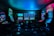 Dimly lit gaming arcade featuring a variety of brightly illuminated slot machines and video gaming screens. Neon signs and colorful displays create a vibrant atmosphere. The arrangement of gaming machines suggests an inviting space for entertainment, with chairs positioned for players.