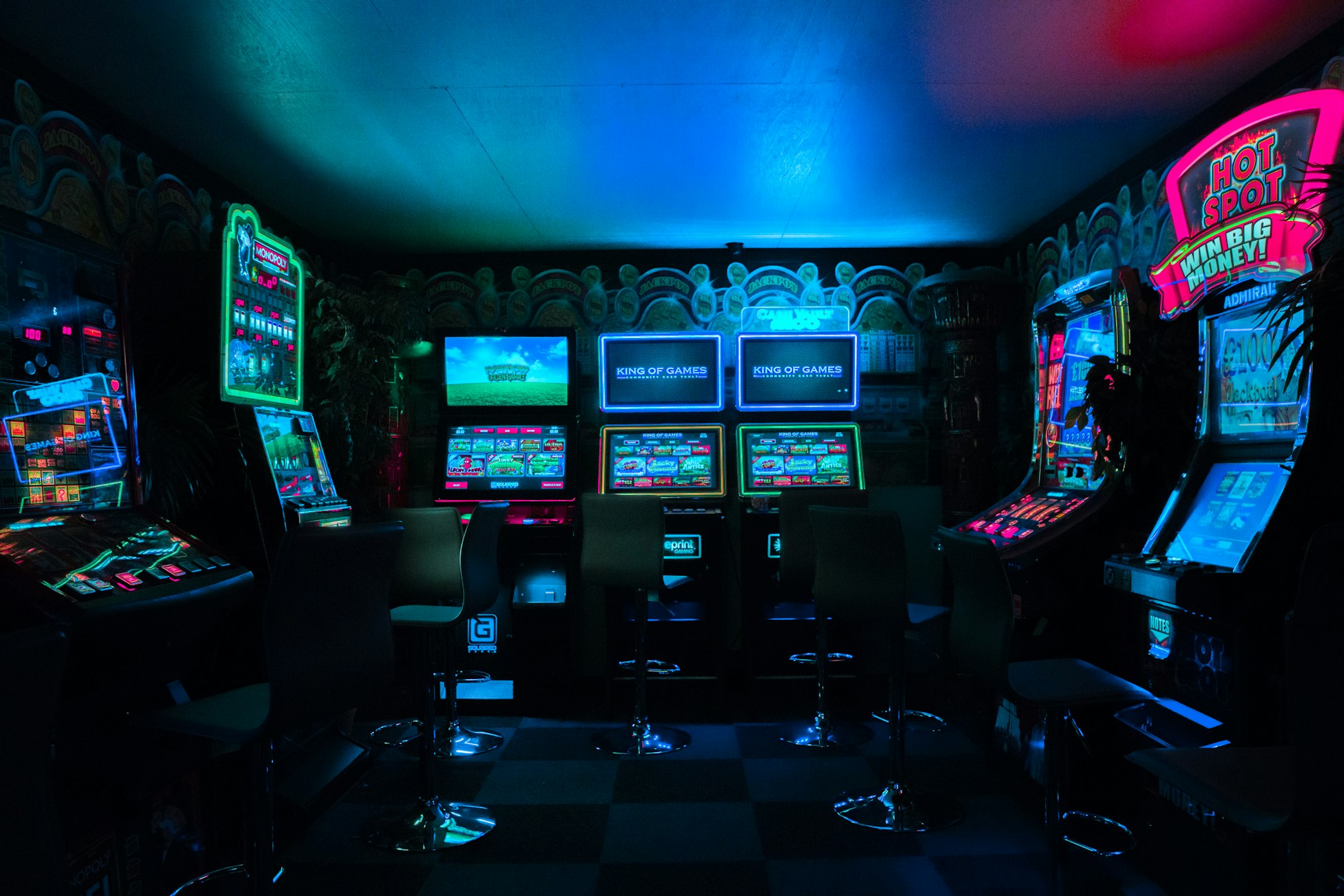 Colorful yet dark photograph of a video arcade, featuring 7 machines. Pink, blue, and green neon light from the machines suffuse the darkened room, lighting up the ceiling.