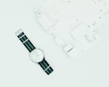 A wristwatch with a white dial and black and blue nylon strap is placed on a light surface next to a white circuit board.