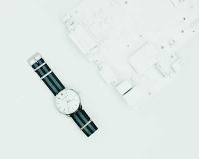 A wristwatch with a white dial and black and blue nylon strap is placed on a light surface next to a white circuit board.