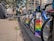 A line of rental bicycles, branded as Citi Bikes, is stationed on an urban sidewalk. The bikes are accompanied by colorful docking stations featuring pride rainbow colors. In the background, there are modern buildings and a few trees. Pedestrians and urban signage add to the cityscape ambiance.