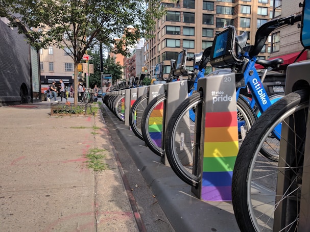 A line of rental bicycles, branded as Citi Bikes, is stationed on an urban sidewalk. The bikes are accompanied by colorful docking stations featuring pride rainbow colors. In the background, there are modern buildings and a few trees. Pedestrians and urban signage add to the cityscape ambiance.