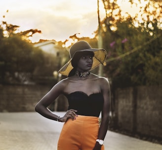 A person stands confidently in a stylish outfit with orange pants and a strapless black top. They wear a wide-brimmed hat that casts a shadow over their face, and a chunky necklace adorns their neck. The background features a blurred, tree-lined street with sunlight filtering through, creating a warm, golden glow.