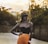 A person stands confidently in a stylish outfit with orange pants and a strapless black top. They wear a wide-brimmed hat that casts a shadow over their face, and a chunky necklace adorns their neck. The background features a blurred, tree-lined street with sunlight filtering through, creating a warm, golden glow.