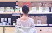 A woman in a white shirt is standing in front of a display shelf filled with various products, including mugs, signs, and postcards. The shelves hold items with inspirational and humorous messages tailored for family members. The environment resembles a gift shop with a focus on family-themed merchandise.