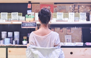 A woman in a white shirt is standing in front of a display shelf filled with various products, including mugs, signs, and postcards. The shelves hold items with inspirational and humorous messages tailored for family members. The environment resembles a gift shop with a focus on family-themed merchandise.