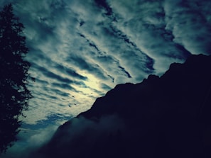 A dramatic landscape with dark, cloudy skies covering the horizon and rugged mountain silhouettes. The scene is accentuated by the faint glow of sunlight attempting to break through the clouds, creating a mystical atmosphere.