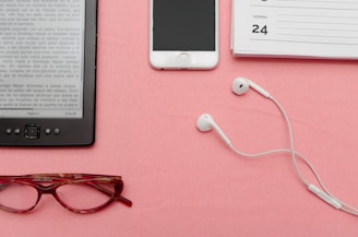 A pair of red reading glasses, an e-reader displaying a page in Spanish, white earphones, a smartphone, and a planner open to the page showing '24th' are neatly arranged on a pink surface.