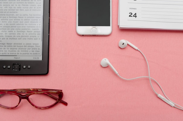 A pair of red reading glasses, an e-reader displaying a page in Spanish, white earphones, a smartphone, and a planner open to the page showing '24th' are neatly arranged on a pink surface.