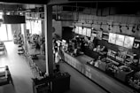 A modern, stylish coffee shop with an industrial design featuring exposed pipes and overhead lighting. The space includes a long wooden table with chairs, various coffee-making equipment, and a countertop where people are seen ordering. Shelves display various products, and light streams in from large windows, illuminating the interior. The atmosphere is lively with several patrons interacting with the baristas.