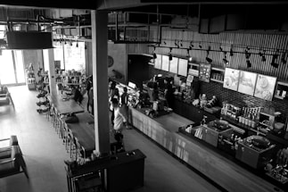 A modern, stylish coffee shop with an industrial design featuring exposed pipes and overhead lighting. The space includes a long wooden table with chairs, various coffee-making equipment, and a countertop where people are seen ordering. Shelves display various products, and light streams in from large windows, illuminating the interior. The atmosphere is lively with several patrons interacting with the baristas.
