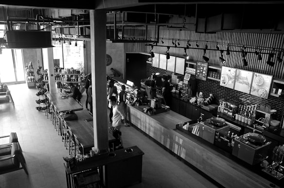 A modern, stylish coffee shop with an industrial design featuring exposed pipes and overhead lighting. The space includes a long wooden table with chairs, various coffee-making equipment, and a countertop where people are seen ordering. Shelves display various products, and light streams in from large windows, illuminating the interior. The atmosphere is lively with several patrons interacting with the baristas.